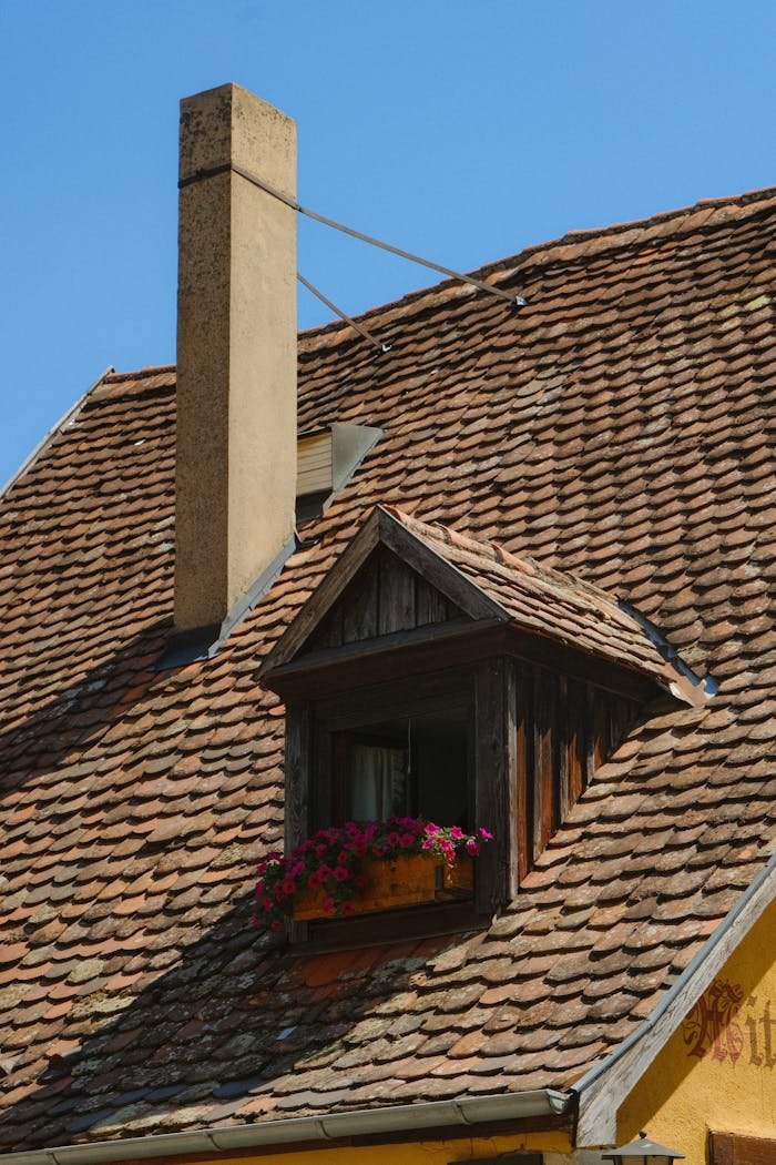 A rustic rooftop with a chimney and window adorned with red flowers against a clear blue sky.